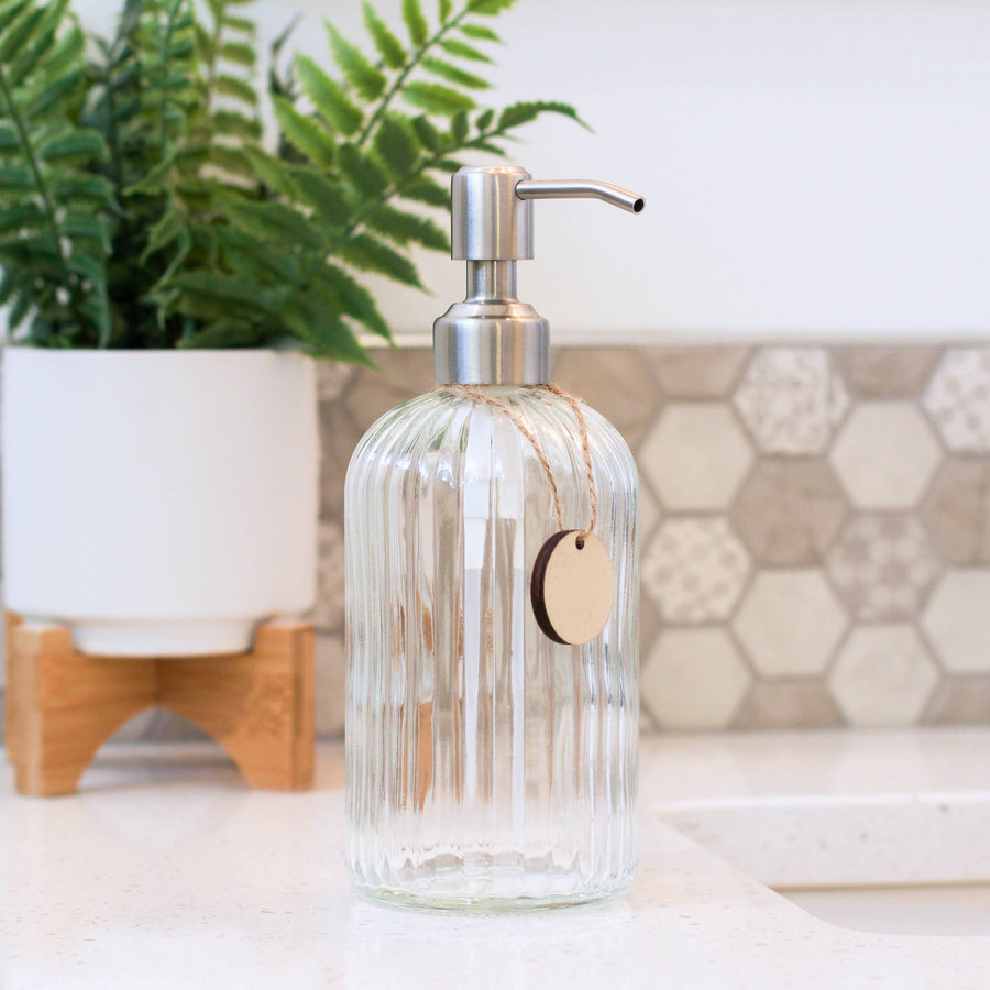 Clear glass soap dispenser with pump on a countertop, plant and textured wall in the background